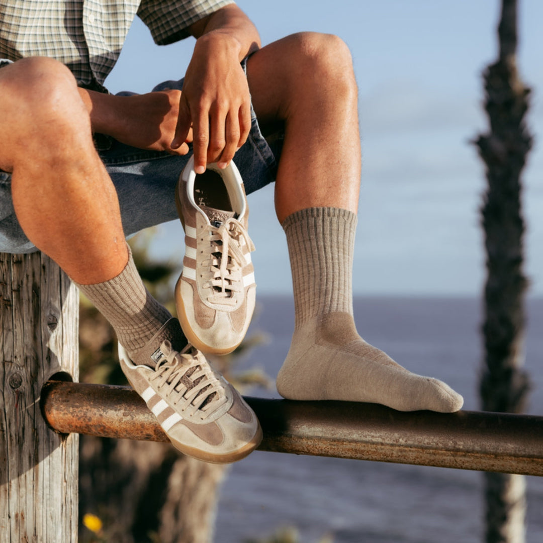 Man sitting on a fence rail holding a sneaker in his hand and wearing tan merino wool crew socks