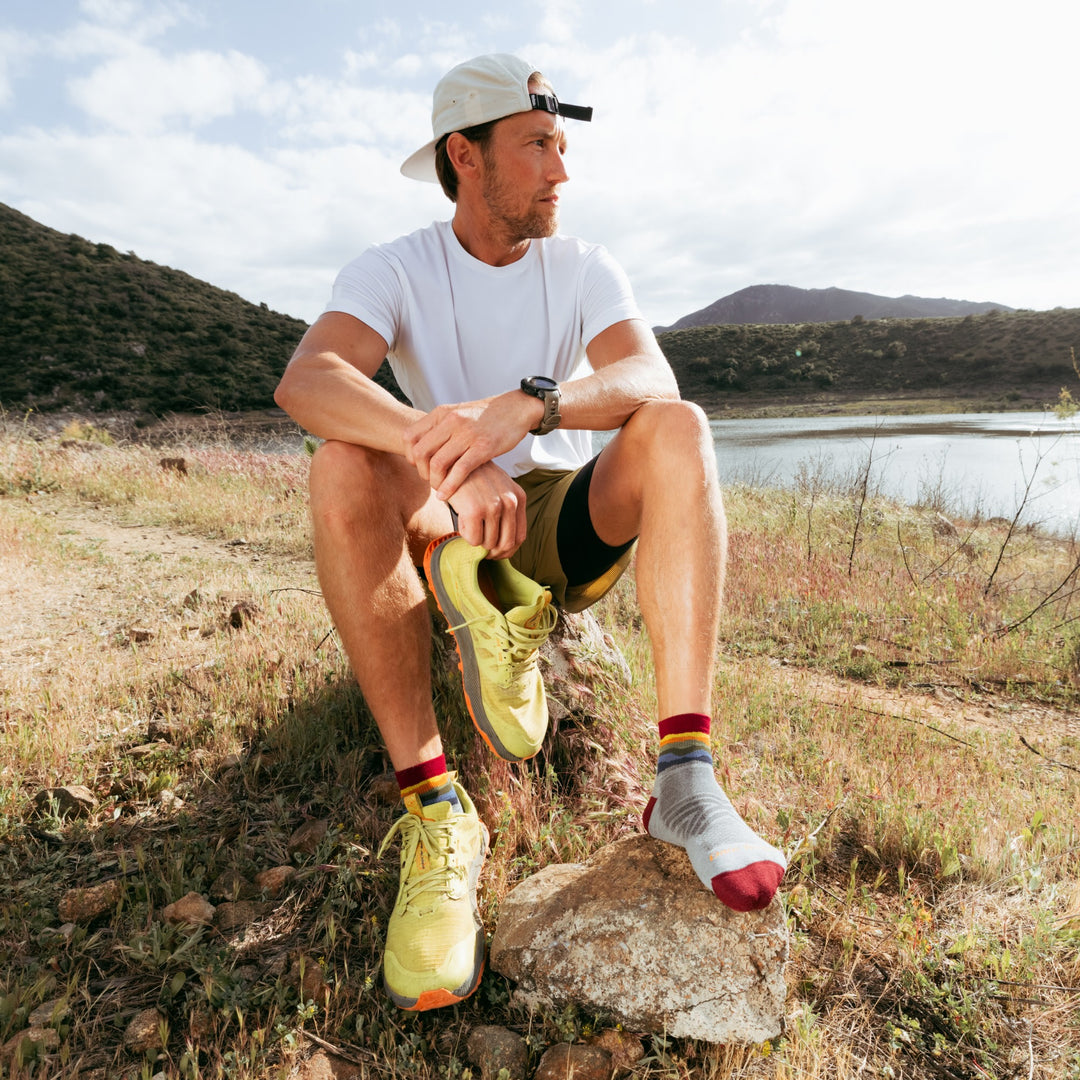 Model sitting on rock next to water holding one shoe in hand wearing the 1040 quarter running sock in skyracer Gray colorway