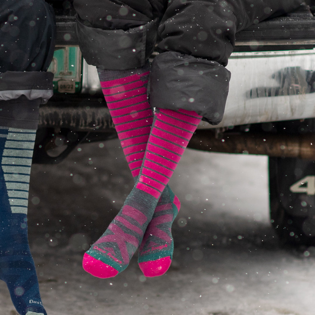 Model sitting in car trunk while it's snowing wearing women's edge over-the-calf snow sock in teal and pink