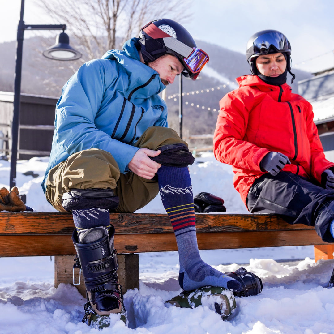 model sitting outside on bench with one ski boot off wearing the 8052 steep lines over the calf ski and snowboard sock in eclipse