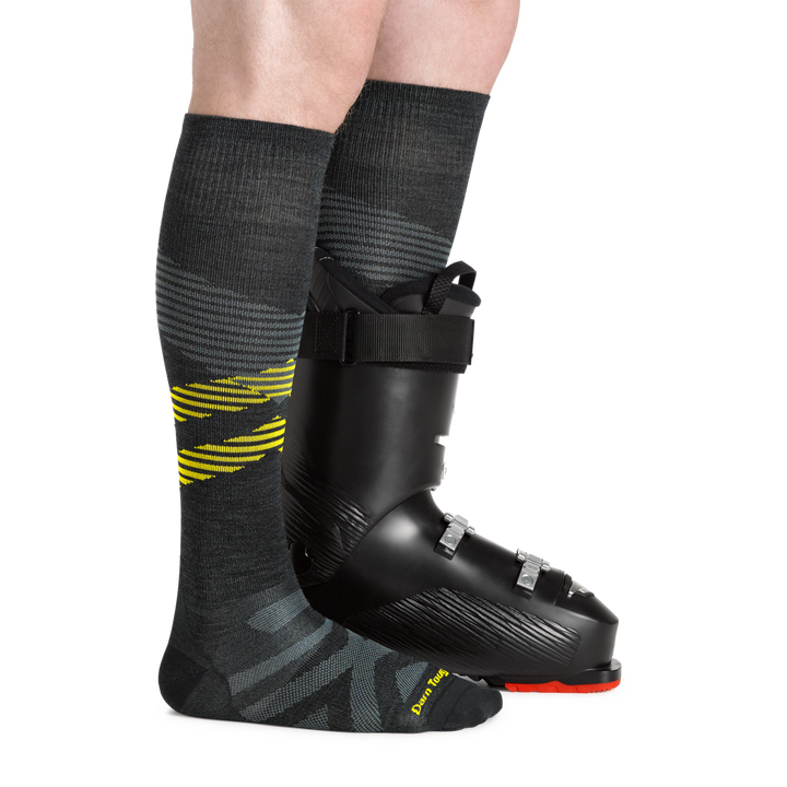 Side studio shot of model wearing men's pennant RFL over-the-calf snow sock in carbon with black snow boot on left foot