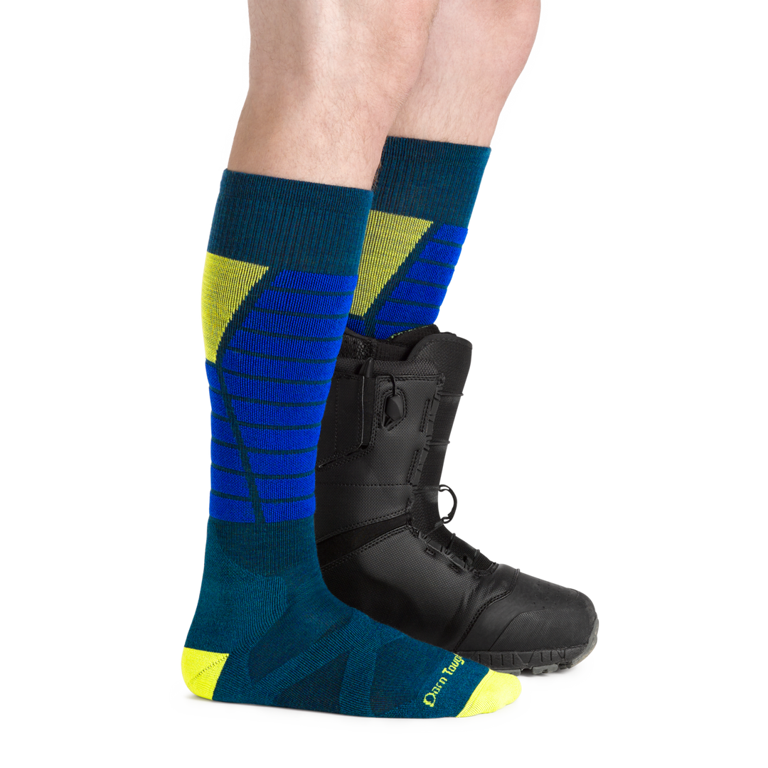 Man facing to the right, wearing Function X Over the Calf Ski & Snowboard Socks in Dark teal, back foot also wearing a snowboard boot