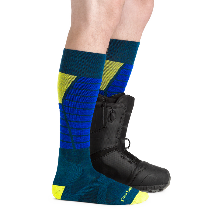 Man facing to the right, wearing Function X Over the Calf Ski & Snowboard Socks in Dark teal, back foot also wearing a snowboard boot