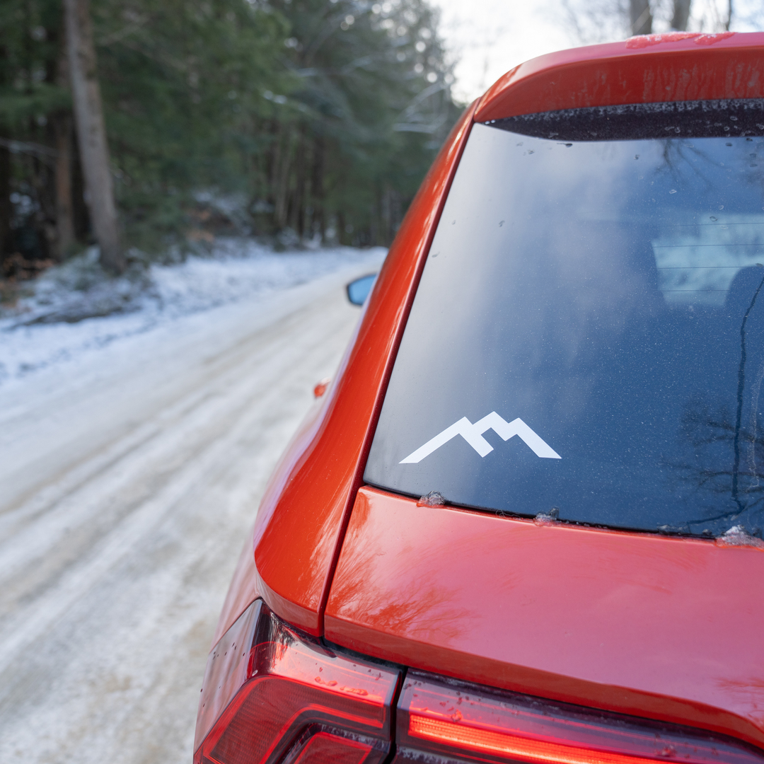 White Darn Tough mountain logo sticker placed on the back window of a red car against snowy road